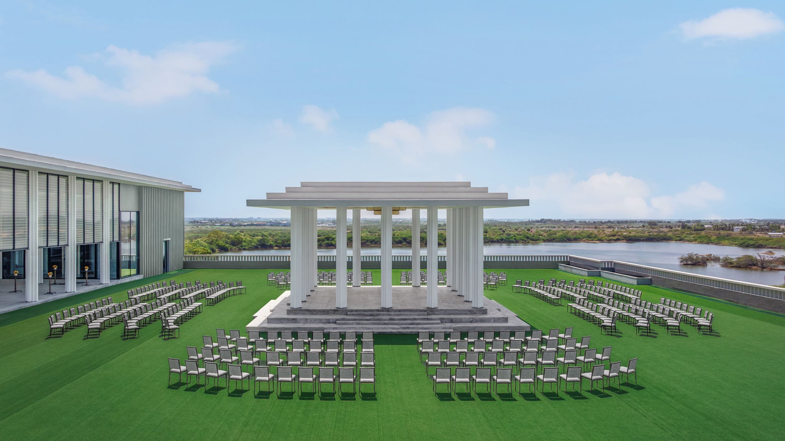 A pavilion with columns, surrounded by rows of chairs, set on a grassy lawn with a view of a body of water and buildings in the distance.