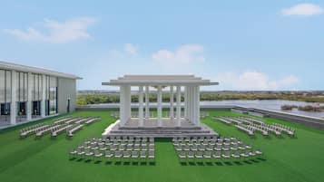 A pavilion with columns, surrounded by rows of chairs, set on a grassy lawn with a view of a body of water and buildings in the distance.