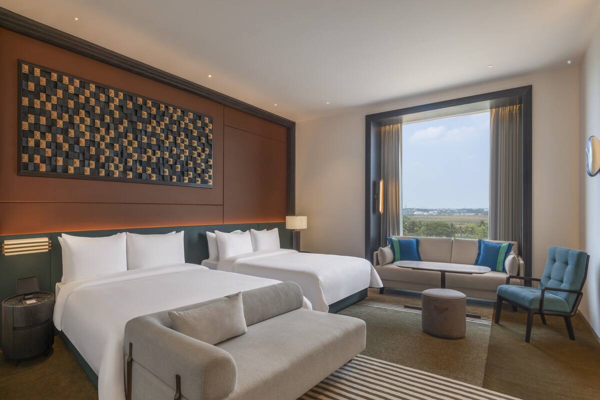 A hotel room with two beds, a sofa, a chair, a small table, a lamp, and a view of the outdoors.