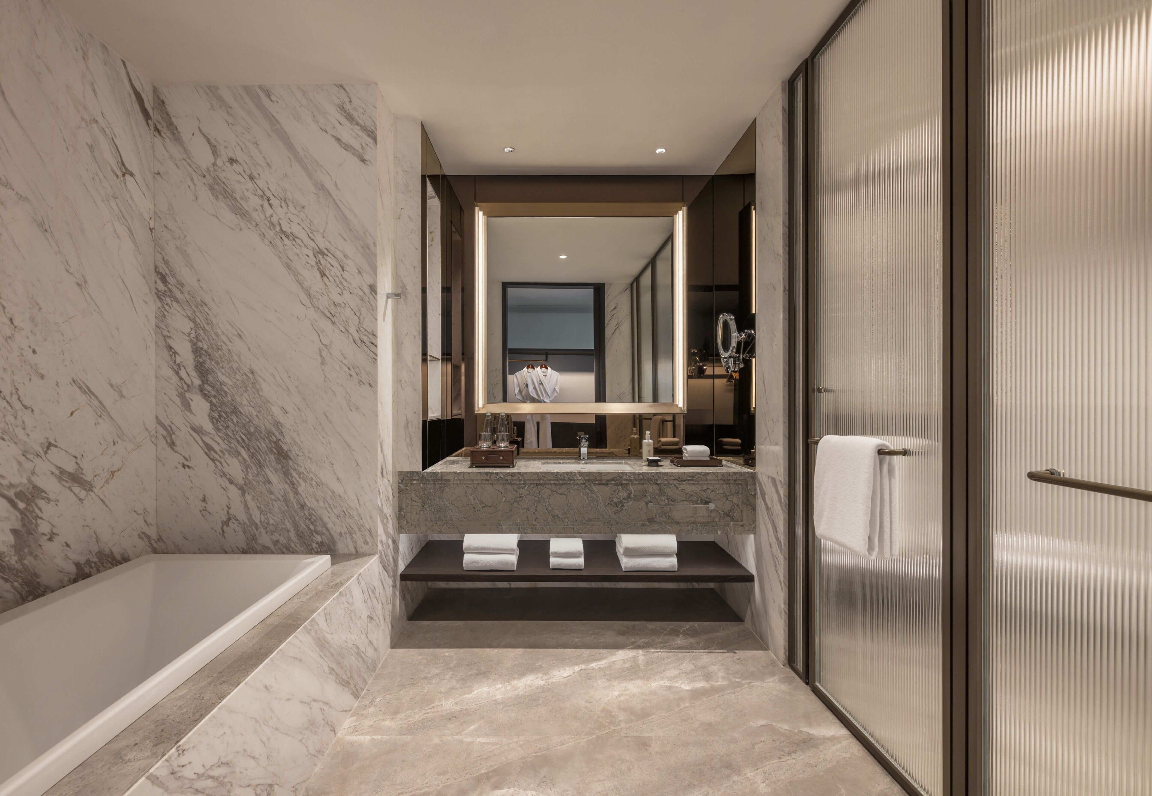 A modern bathroom with a marble wall, a bathtub, a vanity with a mirror, and a shower area.