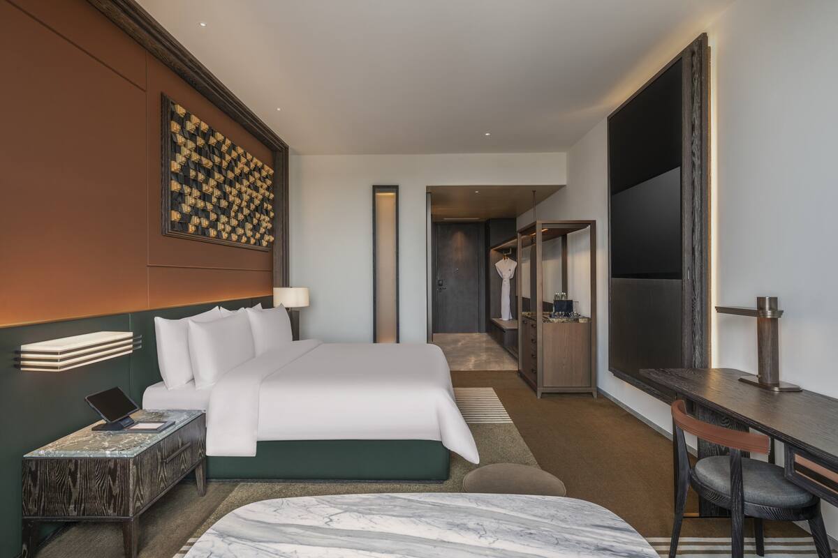 A modern hotel room with a bed, bedside table, desk, chair, and a large flat-screen TV.