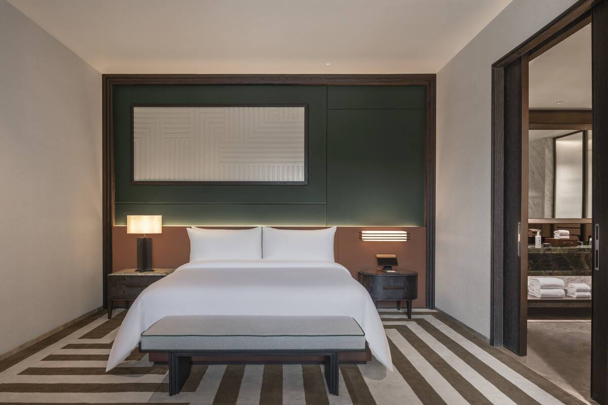 A hotel room with a large bed, bedside tables, a bench, a mirror, and a view into the bathroom.