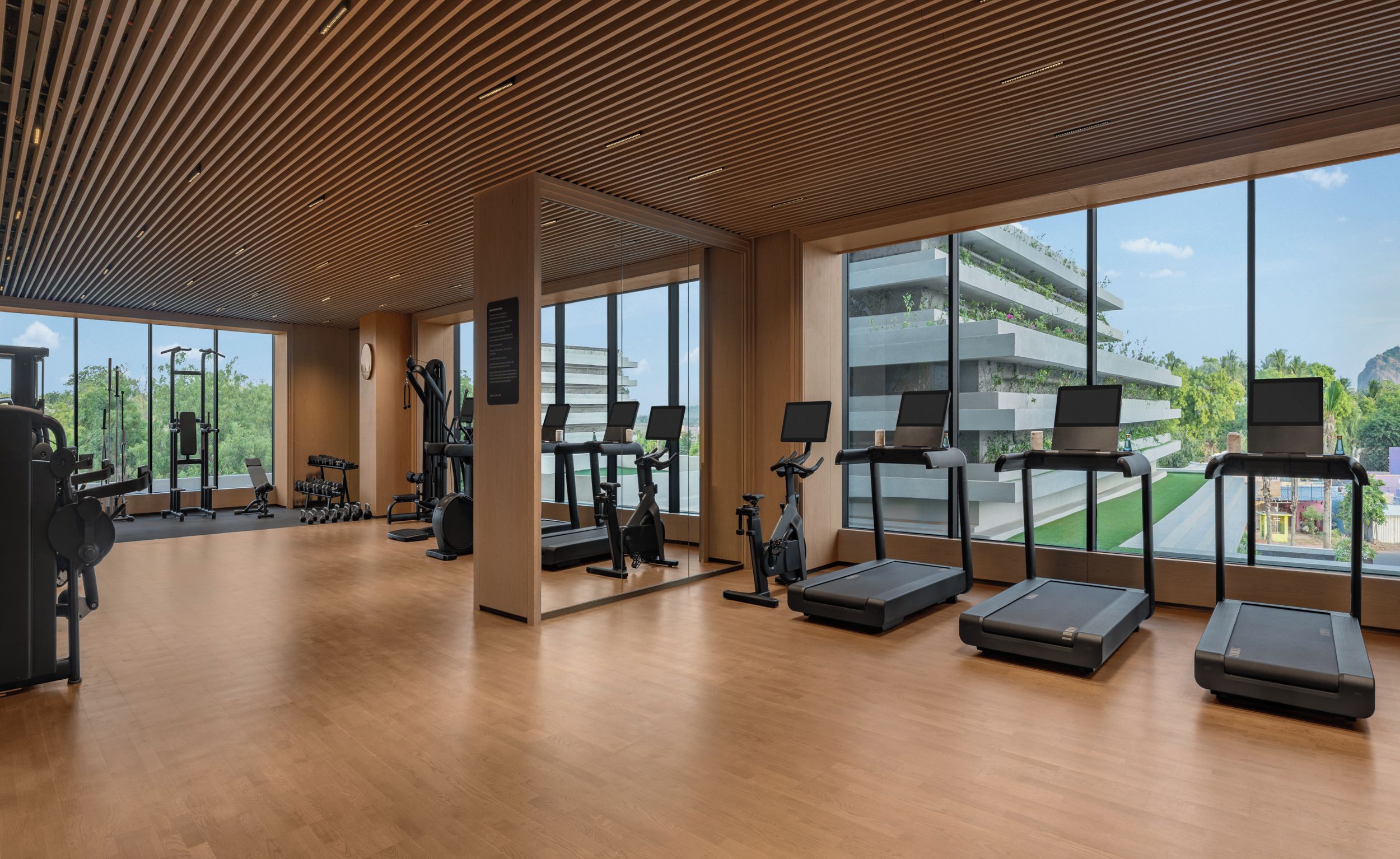 Ein modernes Fitnessstudio mit verschiedenen Trainingsgeräten, großen Fenstern und Blick auf ein Gebäude mit Grünflächen.