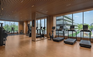 Un gimnasio moderno con diversos equipos de ejercicio, ventanas amplias y vistas a un edificio con vegetación.