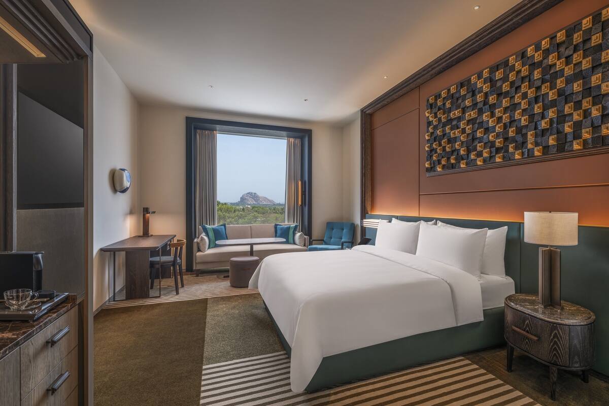 A hotel room with a large bed, a seating area with a sofa and chairs, a dining table, a TV, and a view of the outdoors.