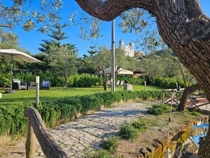 A paved pathway leading to outdoor seating areas with umbrellas, surrounded by lush greenery and a historic building in the background. - A private apartment with an outdoor area for total relaxation (Colli di Fontanelle)