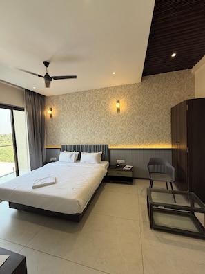 A modern hotel room with a large bed, a ceiling fan, a nightstand with a phone, a chair, and a glass table.