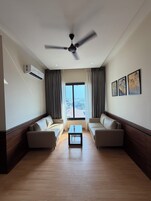 Una sala de estar moderna con ventilador de techo, dos sofás, una mesa de centro y un amplio ventanal con cortinas.