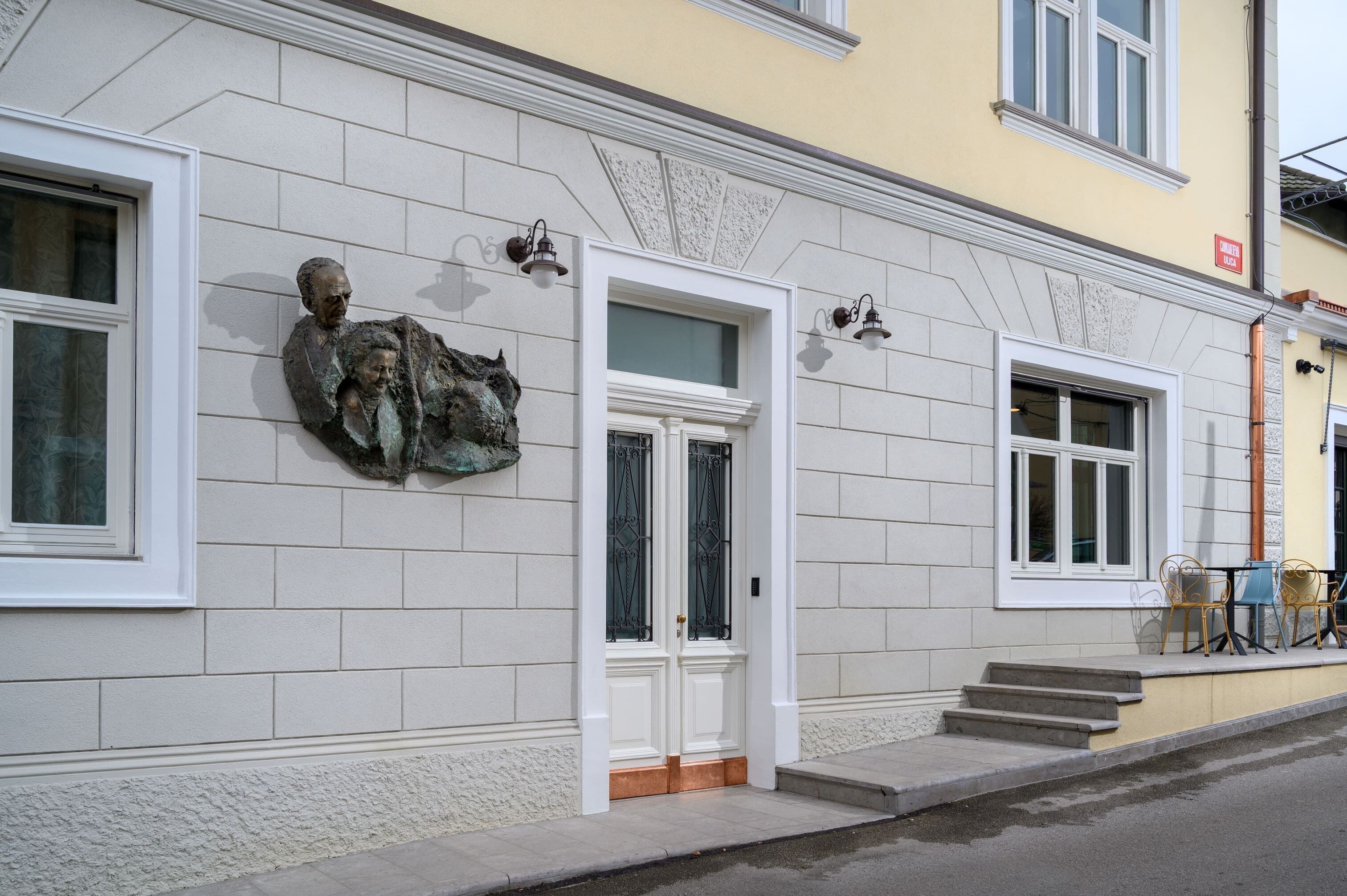 A building facade with a stone wall, a white door, and a small outdoor seating area with metal chairs.