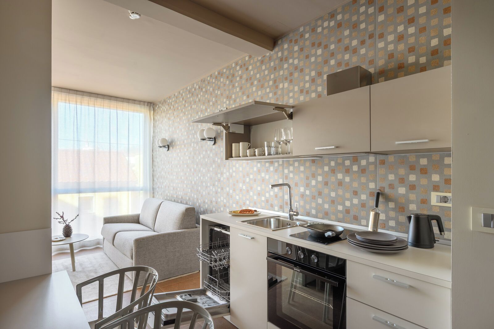Una cocina moderna con horno empotrado, zona de comedor con sofá y ventana con cortinas de gasa.