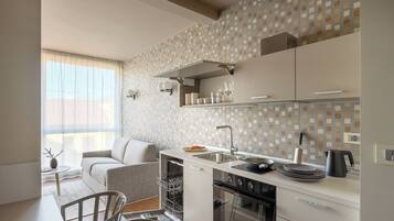 Una cocina moderna con horno empotrado, zona de comedor con sofá y ventana con cortinas de gasa.