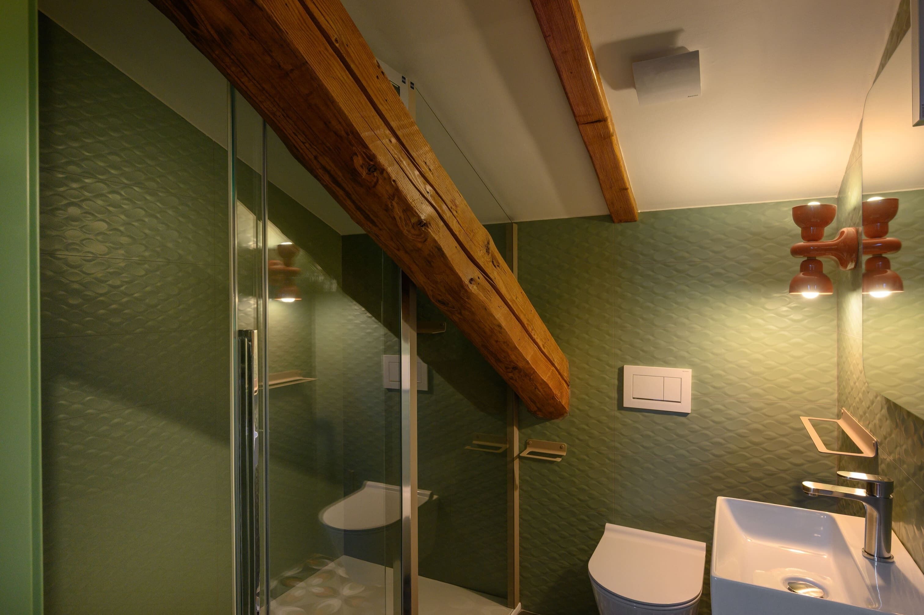 A modern bathroom with a glass shower enclosure, a wooden ceiling beam, and a green textured wall.