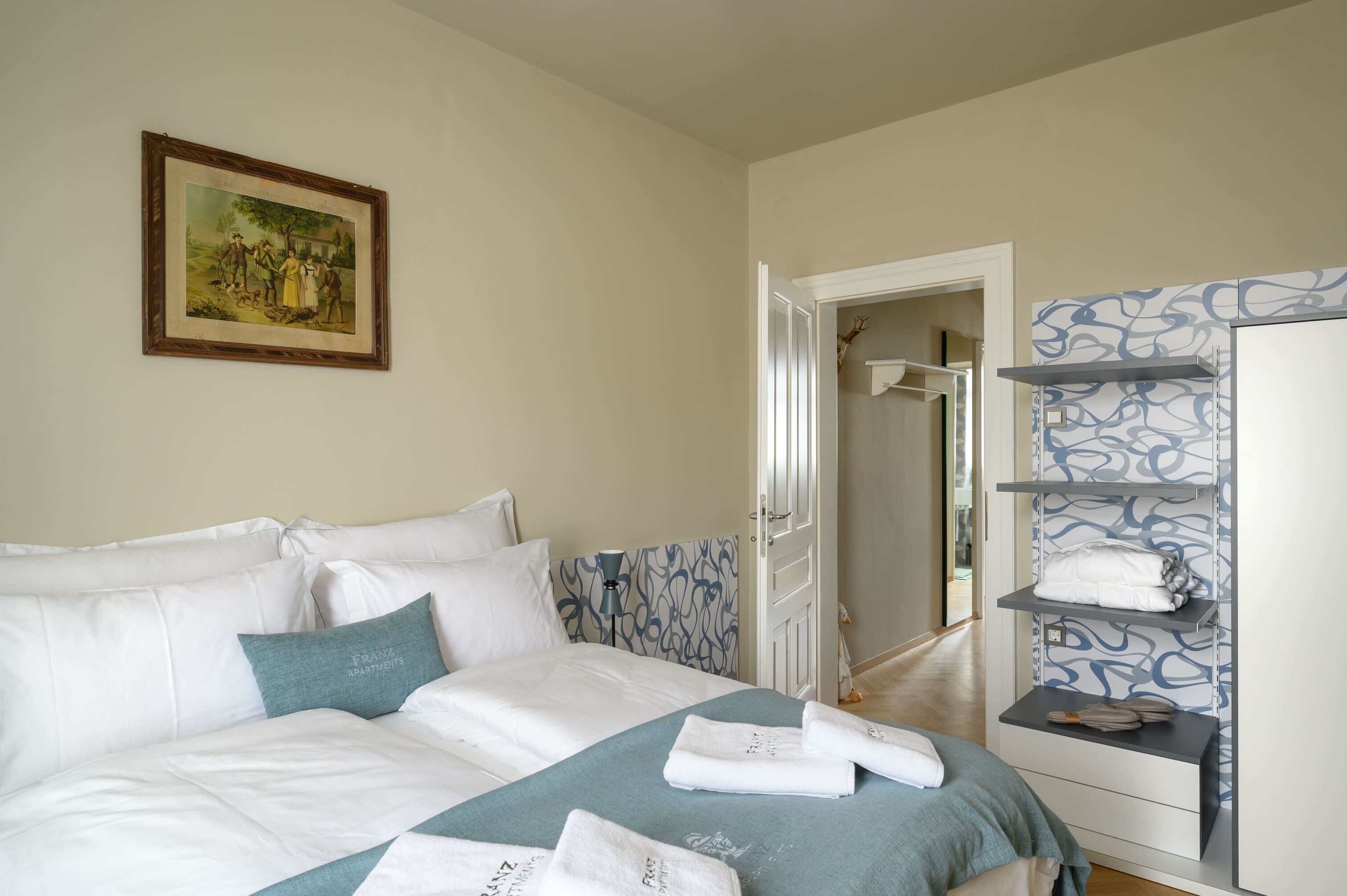 Quarto de hotel com cama, um quadro na parede, um guarda-roupa com prateleiras e uma porta que dá para outro cômodo.