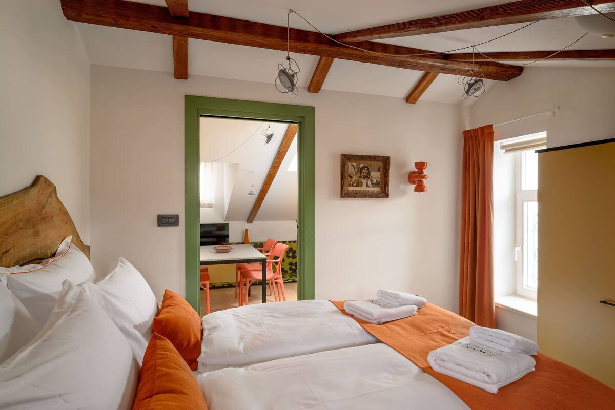 Una habitación con cama, una mesa y sillas, una puerta con marco verde y una ventana con cortinas.