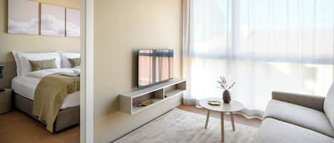 A modern hotel room with a bed, a TV, a small table with a vase, and a large window with sheer curtains.