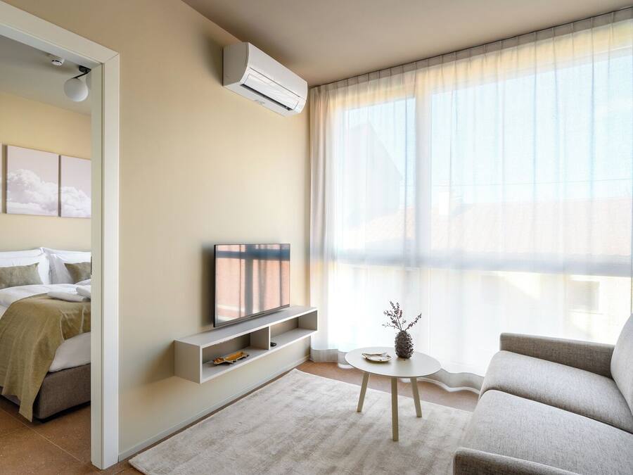 Quarto de hotel moderno com uma cama, televisão, uma mesinha com um vaso e uma janela grande com cortinas leves.