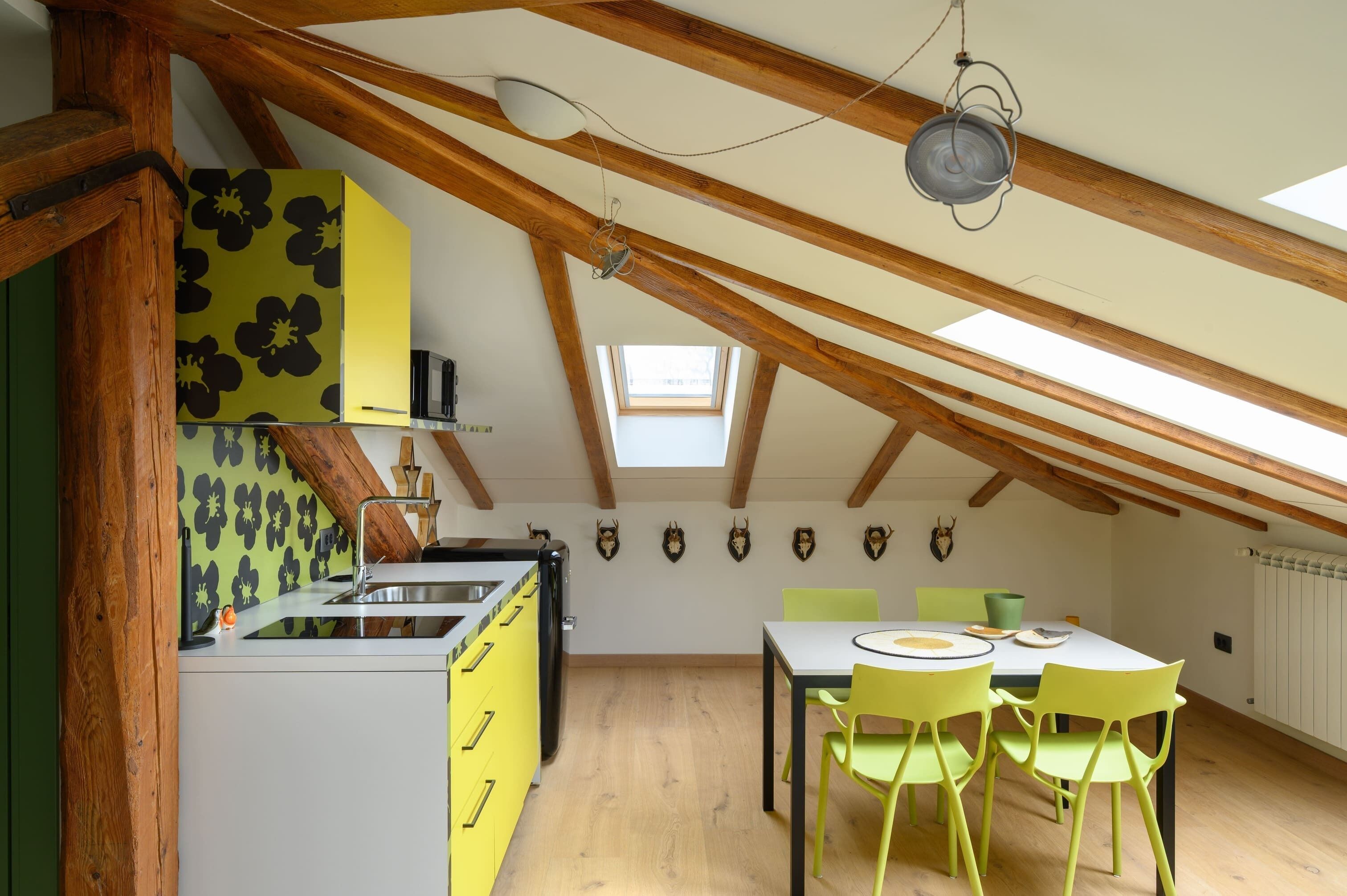 Une cuisine avec un mur recouvert d’un motif floral jaune et noir, des poutres en bois, une verrière et un coin repas avec des chaises vertes.