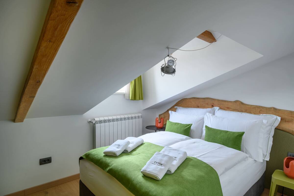 Un dormitorio con techo inclinado, cama con ropa de cama verde y blanca, una mesita con un objeto rojo y una lámpara colgante.
