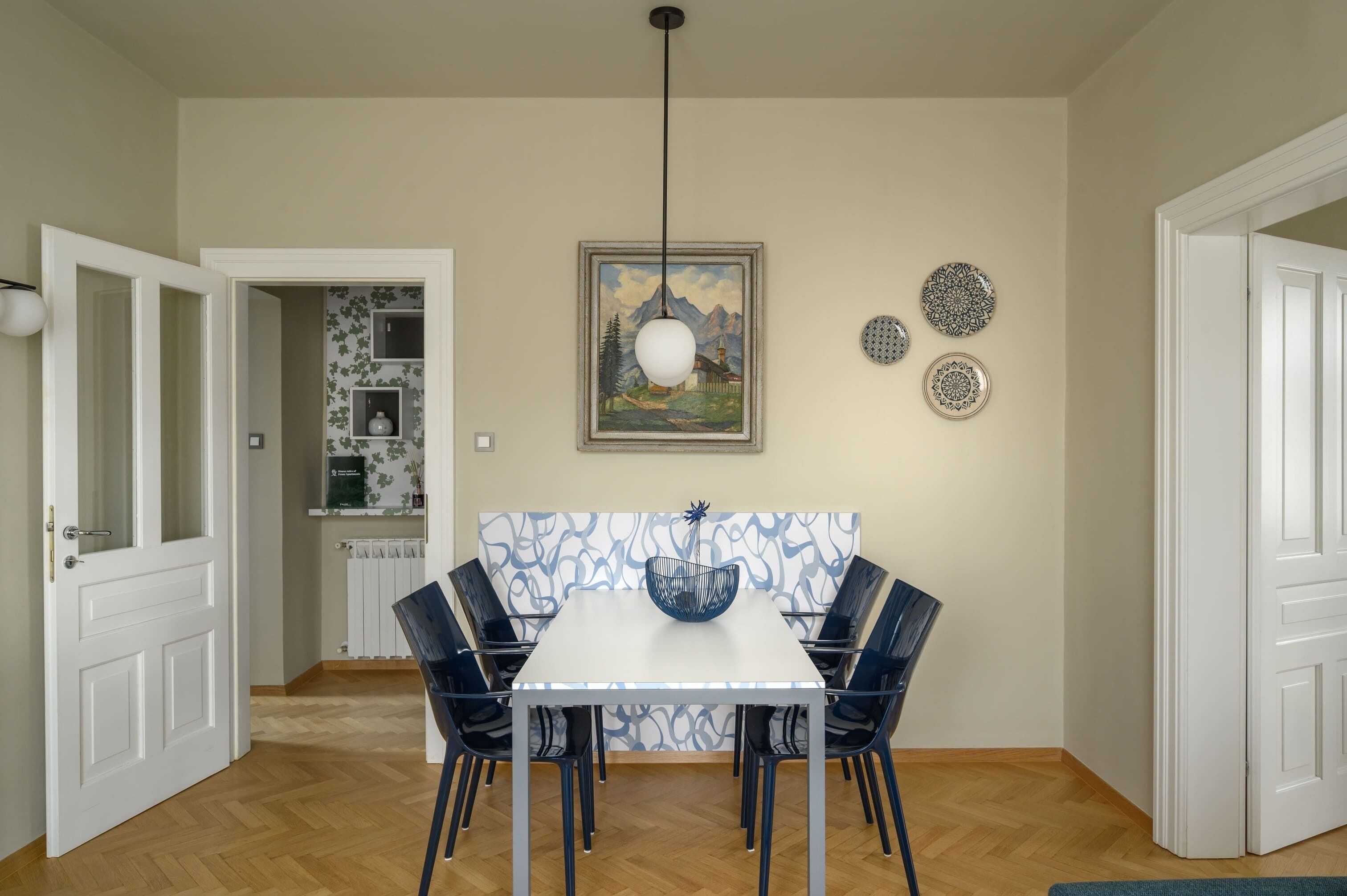 A dining area with a white table and blue chairs, a hanging light fixture, and a framed painting on the wall.