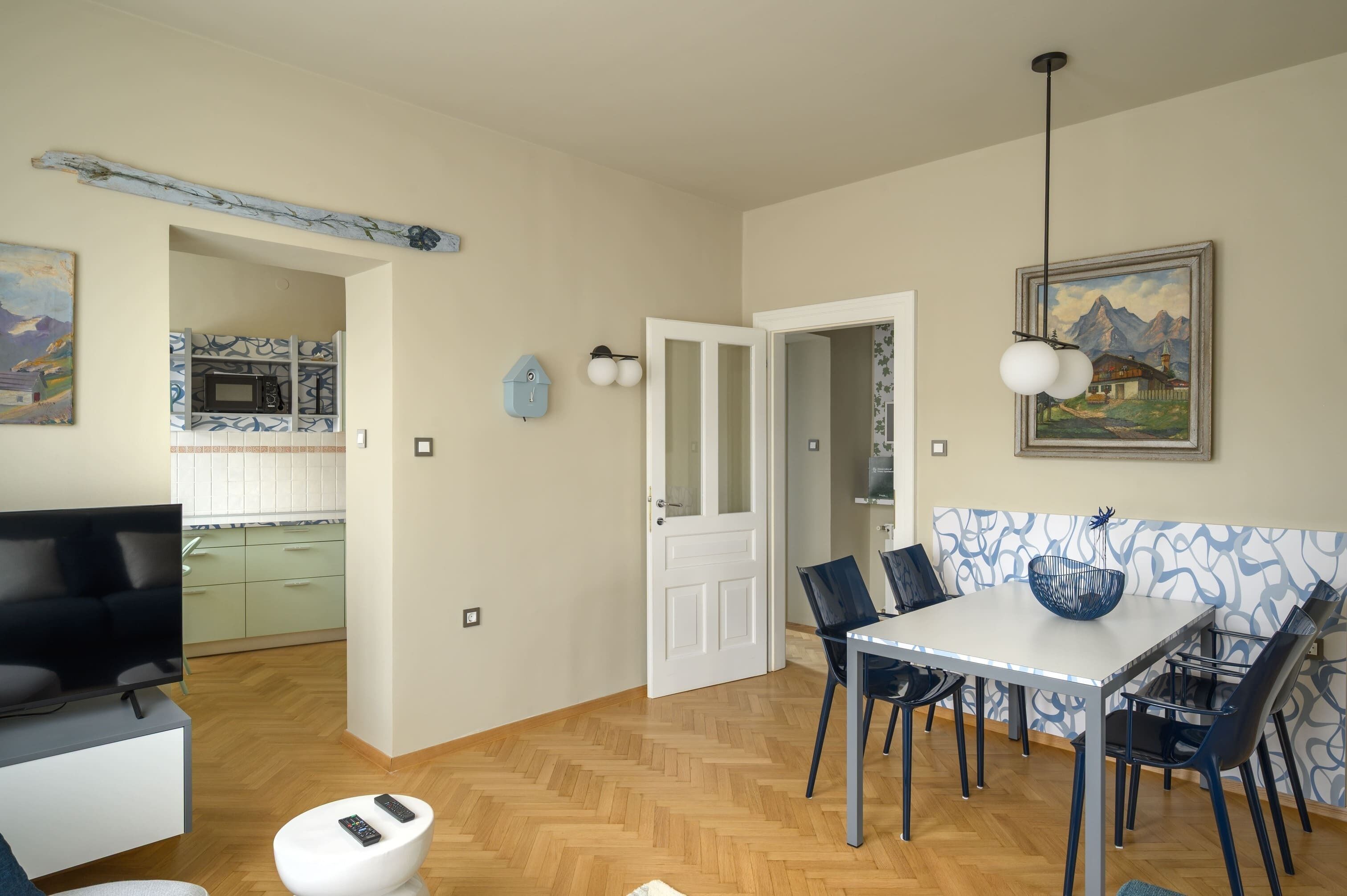 Uma sala moderna com televisão, área de jantar com mesa e cadeiras, cozinha com micro-ondas e uma porta que dá para outro quarto.