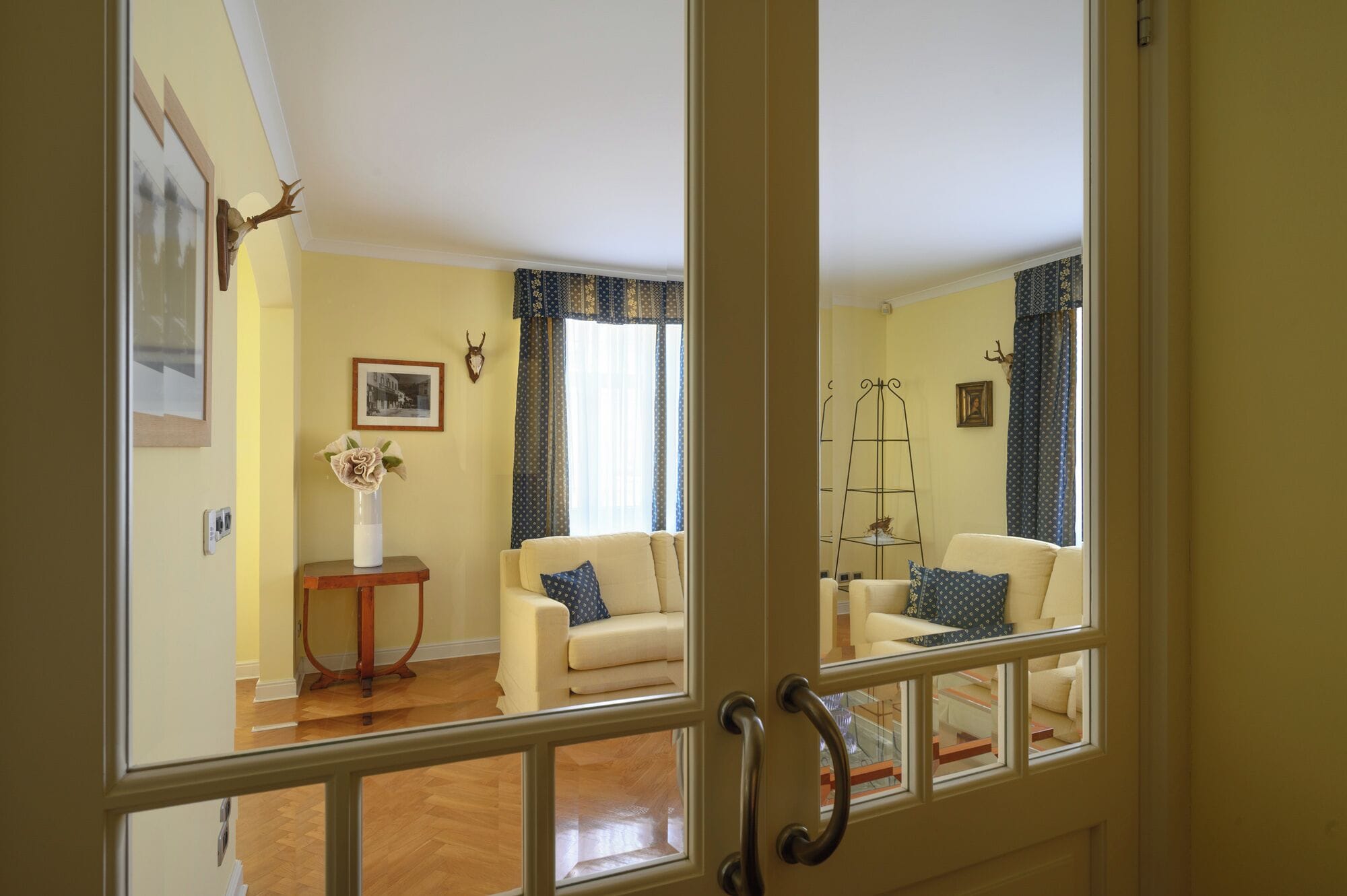 A room with two cream-colored sofas, a small table with a vase, a framed picture, and a decorative antler on the wall.