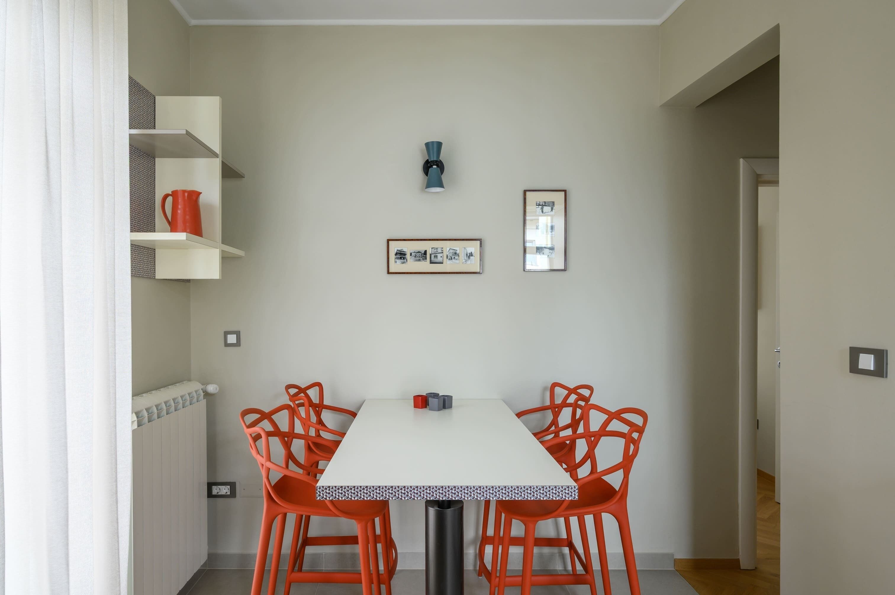 Spiseområde med et hvidt bord og orange stole, en radiator, en blå væglampe og billeder i rammer på væggen.