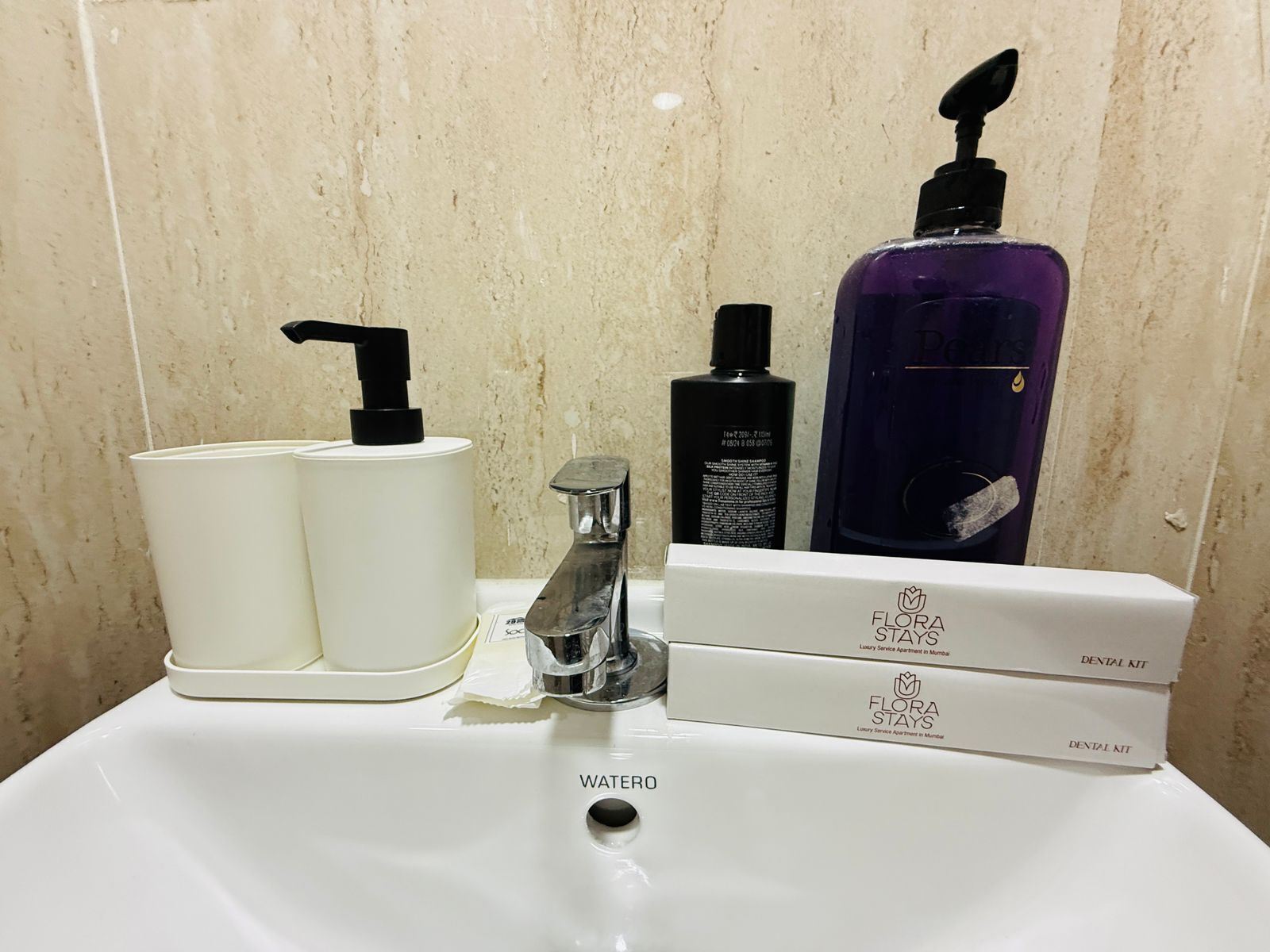 A bathroom sink with a water faucet, a soap dispenser, and two bottles of liquid soap. There are also two boxes labeled "Flora Stays" and "Dental Kit" on the sink.