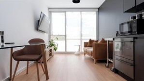 Una cocina moderna con un televisor de pantalla plana, un comedor con una silla, un sofá y un amplio ventanal que proporciona luz natural.