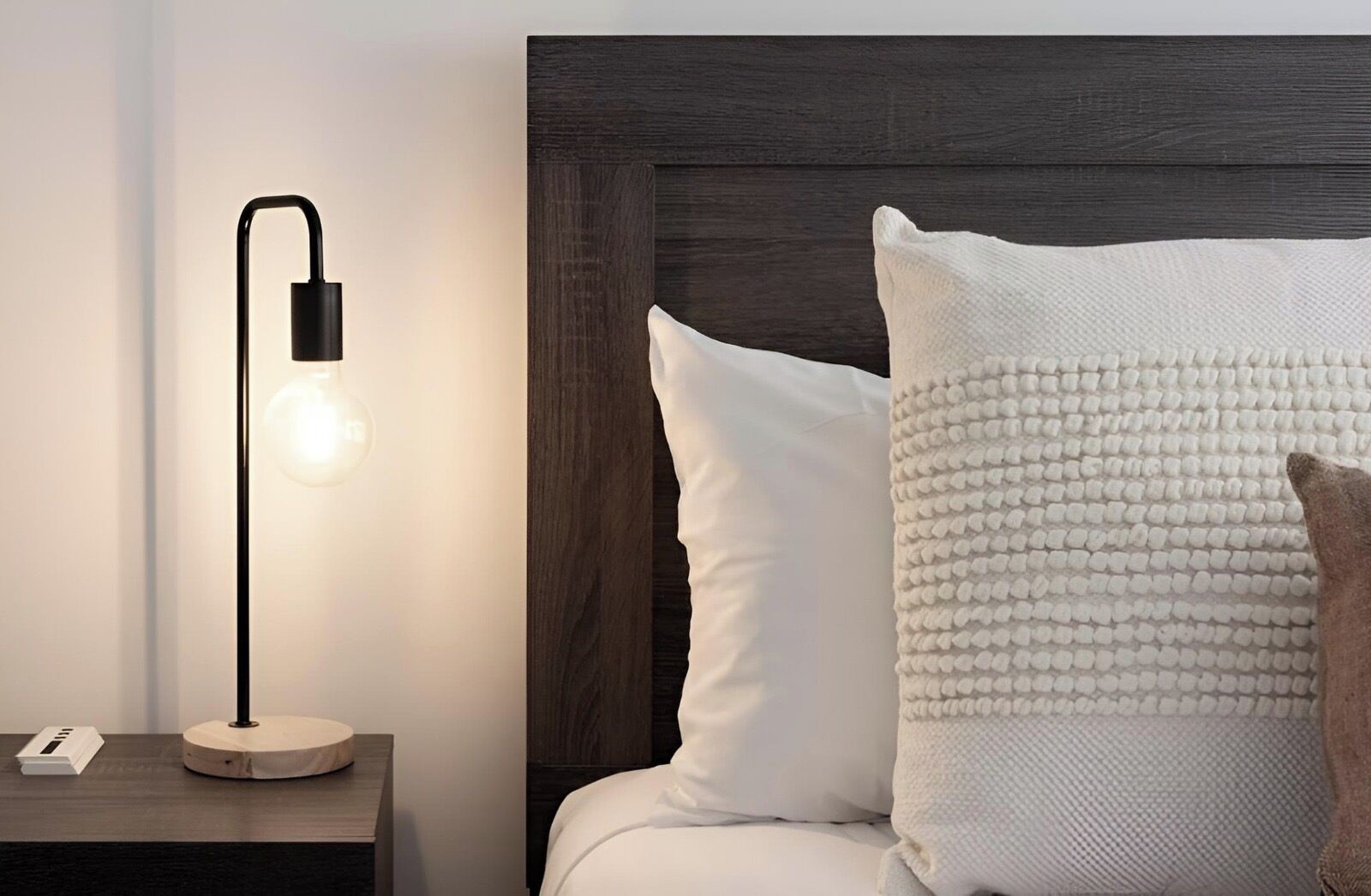 A neatly made bed with white and beige pillows, a dark wooden headboard, and a modern floor lamp with a light bulb on a bedside table.