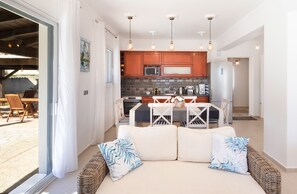 A modern kitchen with wooden cabinets, a dining area with a white table and chairs, and a seating area with cushions.