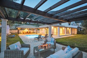 A covered outdoor patio with wicker furniture, a pool, and a house in the background.
