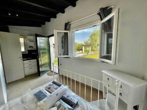 A room with a kitchen, a window with an open view, a white dresser, and a bed with a plastic cover.