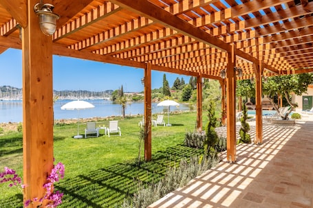 A wooden pergola with a view of a lake and boats, outdoor seating, and a pool area.