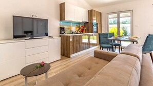 Una sala de estar moderna con un sofá, una mesita con una rosa, un televisor y una zona de cocina con armarios y una ventana.