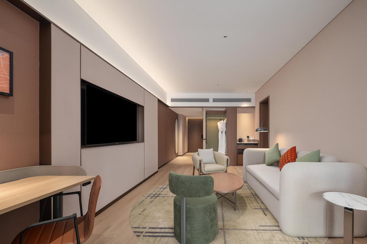 Una habitación de hotel moderna con una televisión de pantalla plana, un sofá, una pequeña mesa redonda, un escritorio con sillas y un vestidor.