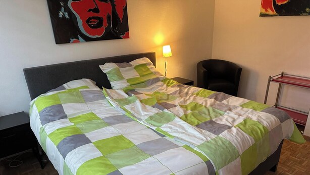 A neatly made bed with a green and white checkered bedspread, a bedside lamp, a chair, and a small table with a lamp. - Room-in the Heart of the City of Salzbur (Salzburg)