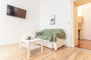 A room with a white sofa, a small table with a plant, a framed picture on the wall, and a TV mounted on the wall. - Simple 1BR Apartment Long Stay Belvedere (Vienna)