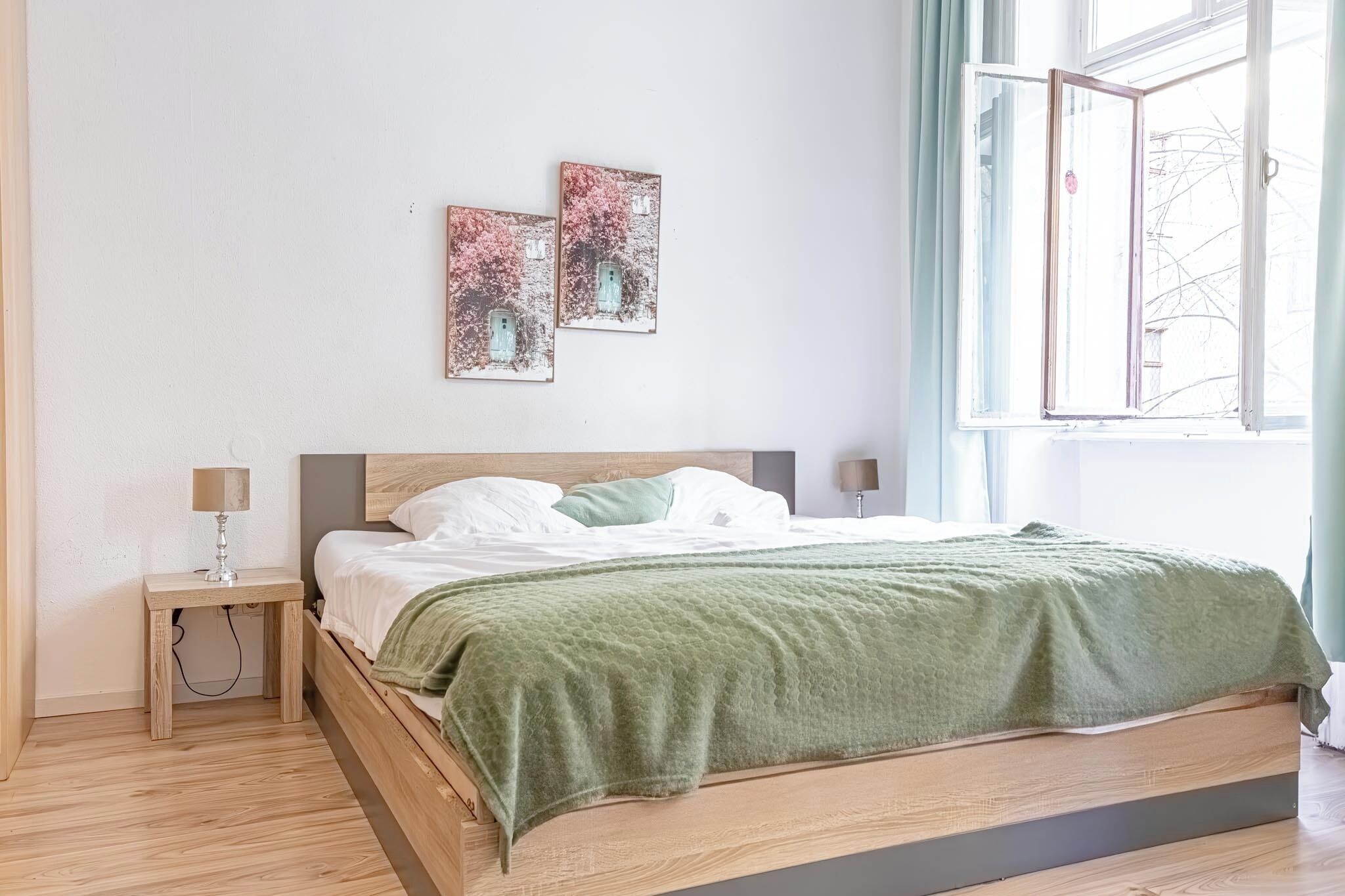 A bedroom with a wooden bed, a green blanket, two bedside tables with lamps, and artwork on the wall.