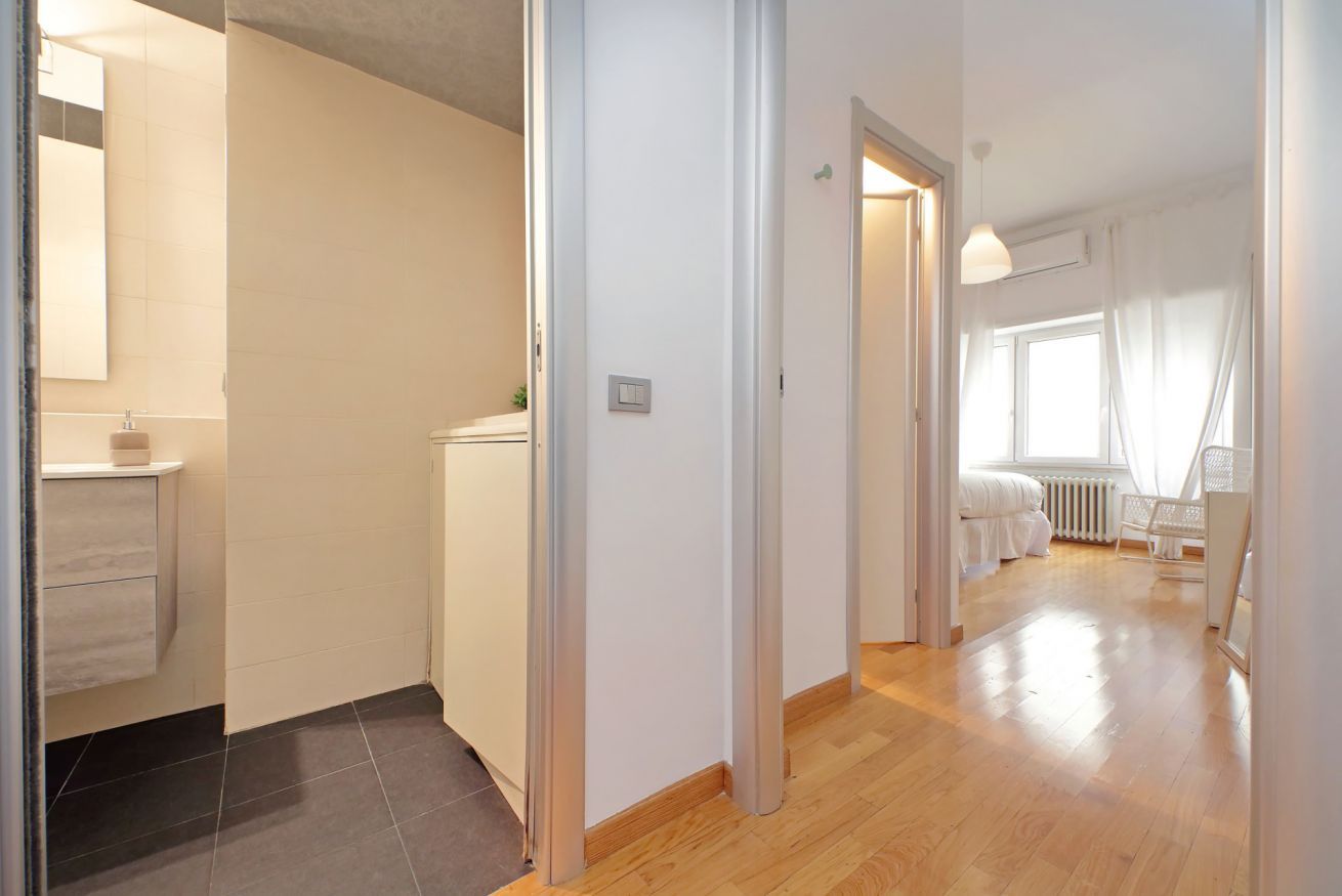 A hallway with a bathroom on the left, a white door leading to a bedroom, and a window with curtains.