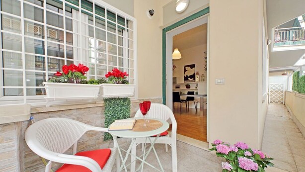Ein gemütlicher Sitzbereich im Freien mit weißen Korbstühlen und einem kleinen runden Tisch, ein Fenster mit roten Blumen und ein Blumentopf mit rosa Blüten.