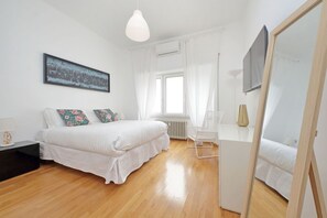 Ein Schlafzimmer mit Bett, Nachttisch, Sessel, Fenster mit Vorhängen, Heizkörper und Spiegel.