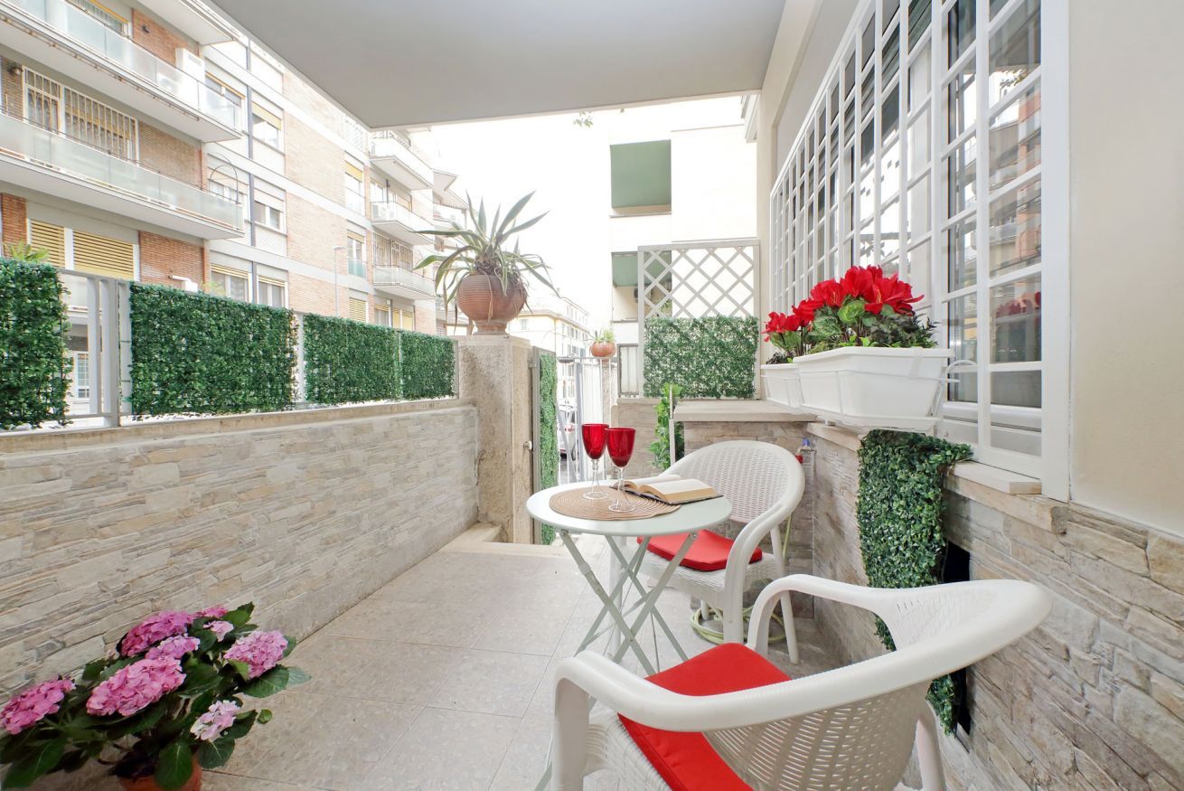 Ein Balkon mit weißen Korbstühlen, einem Tisch mit roten Kissen und Topfpflanzen.