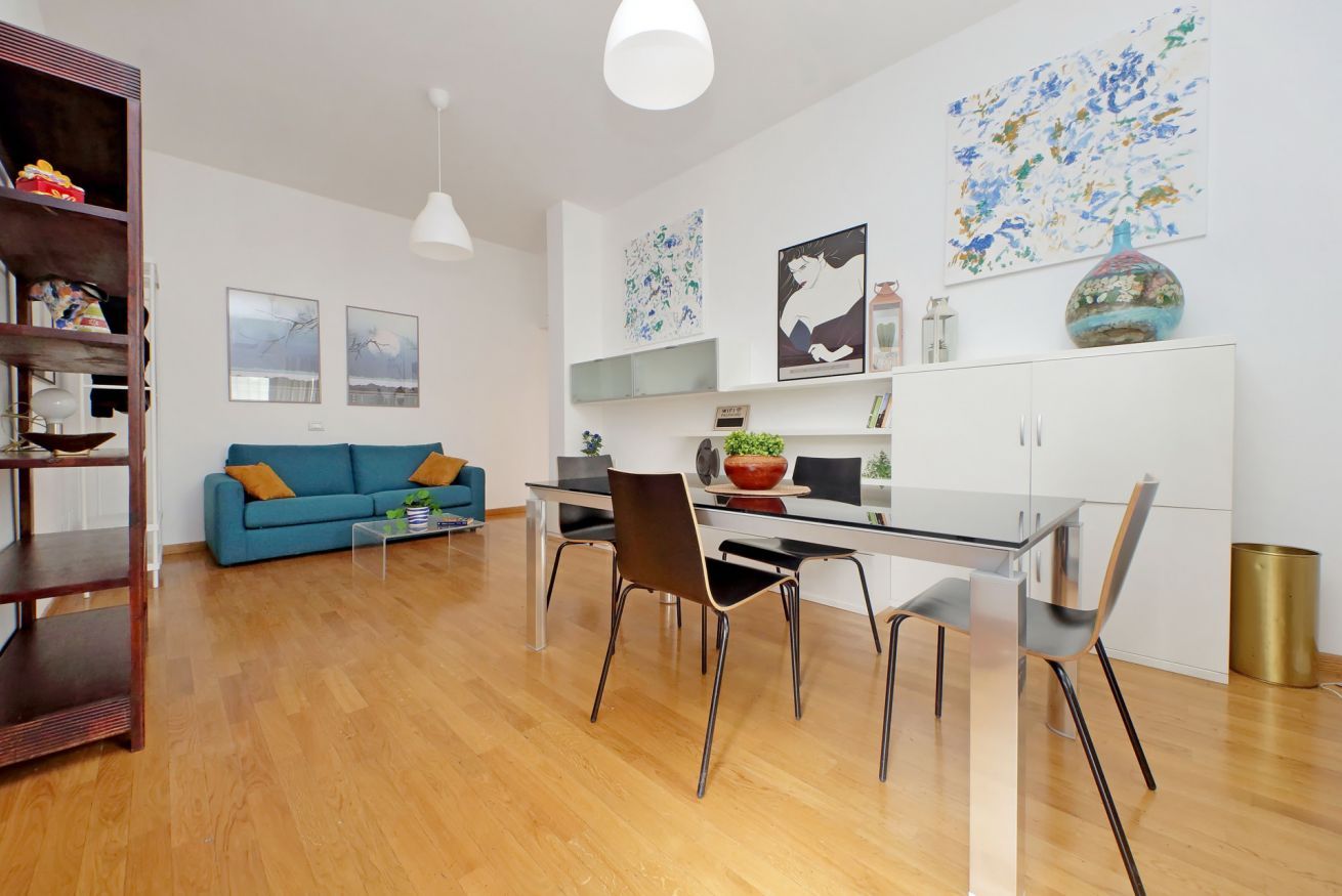 Ein modernes Wohnzimmer mit einem blauen Sofa, einem Esstisch mit Stühlen und einer Küchenzeile mit Schränken und einer Vase.