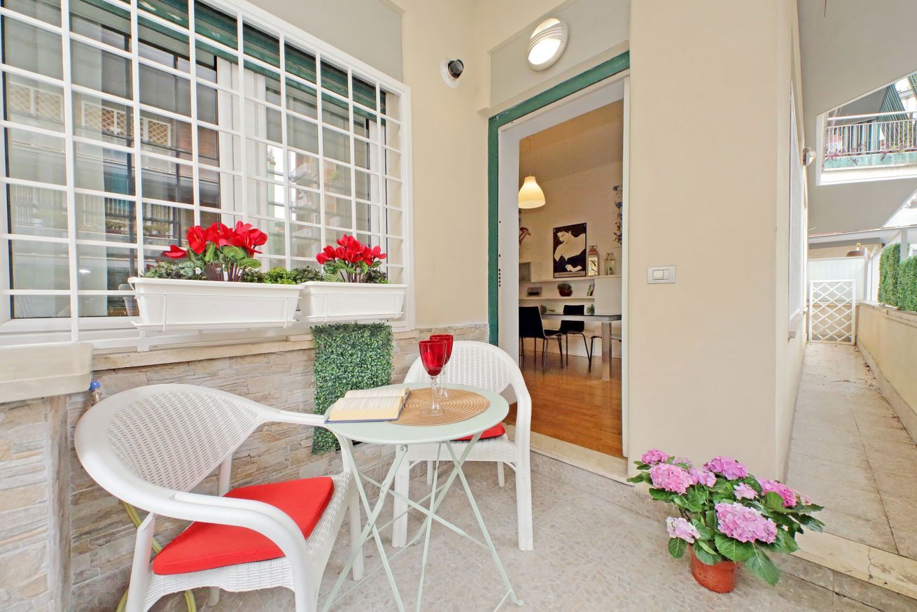 A cozy outdoor seating area with white wicker chairs and a small round table, a window with red flowers, and a potted plant with pink flowers.