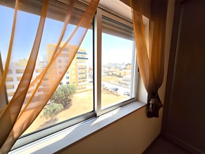 Ein Fenster mit Vorhängen, Blick auf ein Wohngebiet mit Gebäuden und einer unbefestigten Straße.