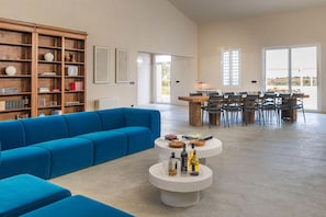 Ein geräumiger Wohnbereich mit einem blauen Ecksofa, einer Holzbibliothek, einem runden Couchtisch mit Flaschen sowie einem Essbereich mit Stühlen und Tisch.