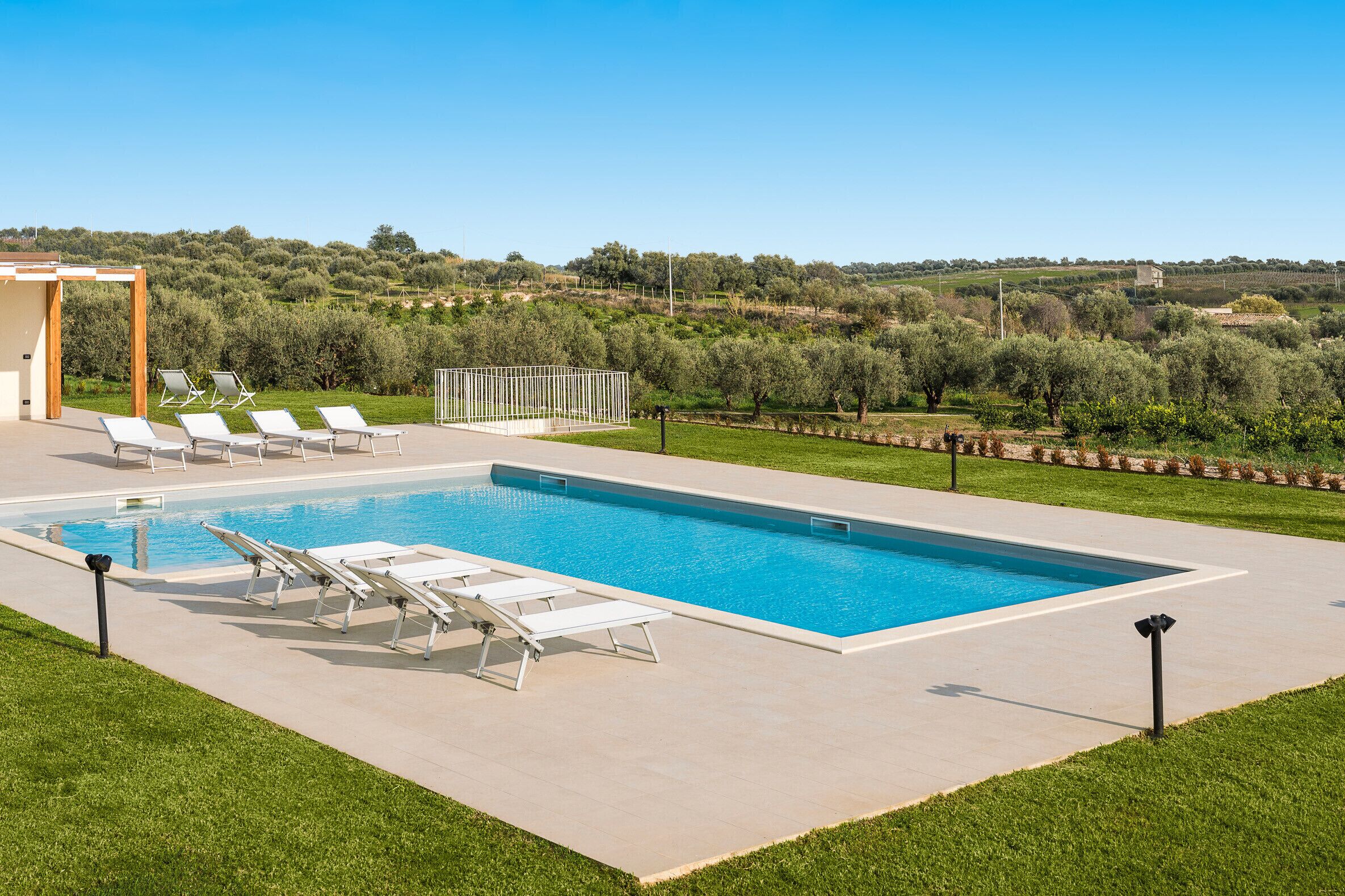 A rectangular swimming pool with a clear blue water surface, surrounded by a light-colored deck. There are several white lounge chairs aligned along the pool's edge, and a small wooden structure is visible in the background.