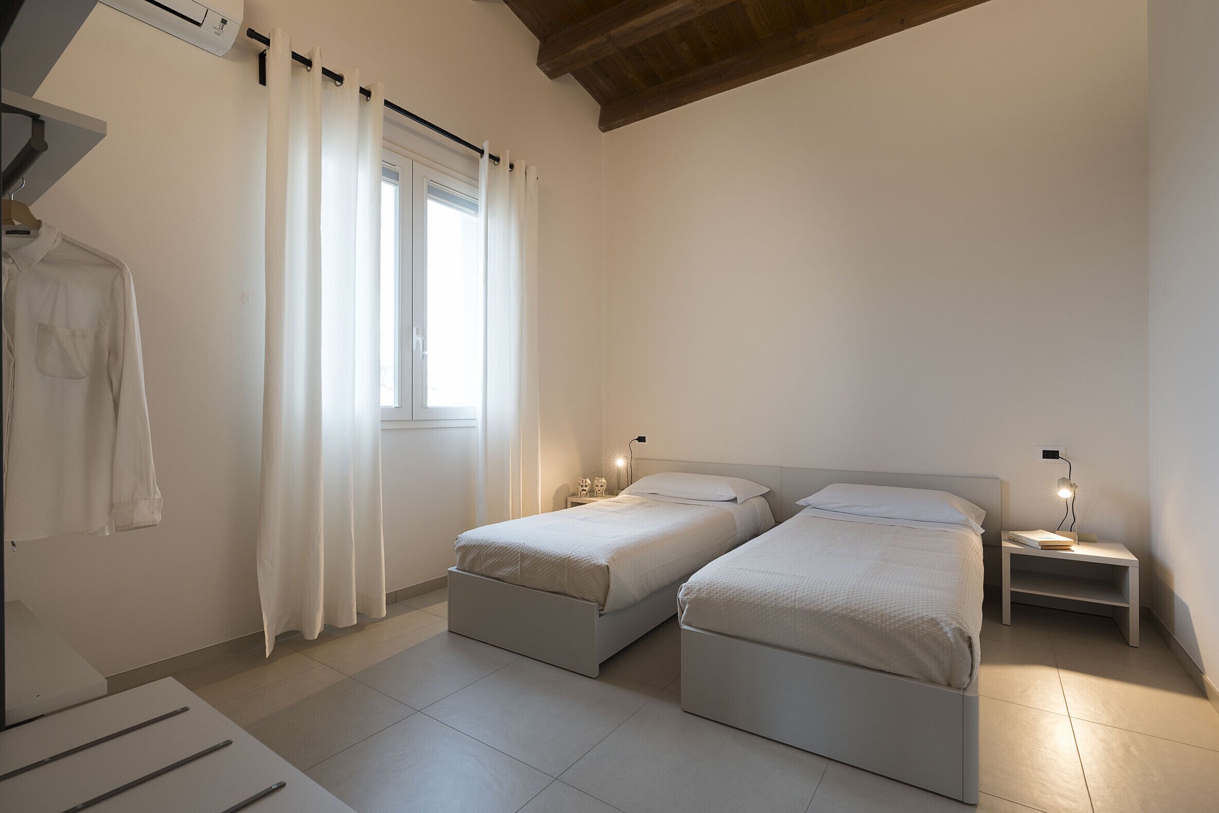 Quarto com duas camas, uma mesa de cabeceira com abajur, uma janela com cortinas e um roupão pendurado.