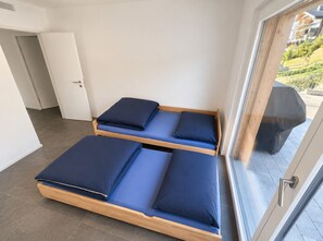 Ein Zimmer mit zwei Holzbettgestellen, blauer Bettwäsche, einer weißen Tür und Blick auf einen Balkon mit Gartenmöbeln.