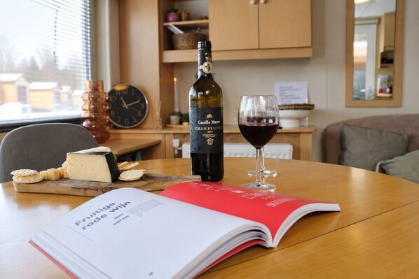 Una botella de vino Castille Mayor, una copa de vino tinto, queso, galletas y un libro sobre una mesa en una habitación con un reloj y armarios al fondo.